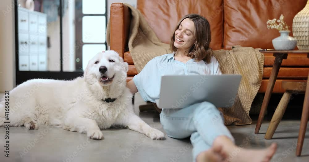 Young woman works on a laptop while sitting with her adorable white dog on the floor at cozy home