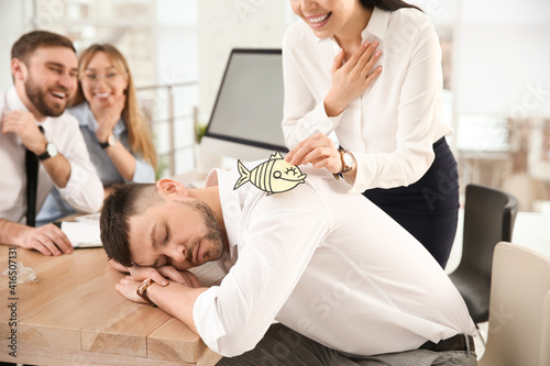 Young woman sticking paper fish to colleague's back while he sleeping in office, closeup. Funny joke