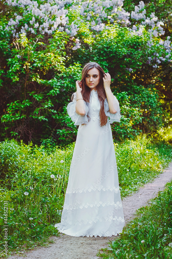 Portrait of young attractive woman in long white wedding dress in spring garden with blooming trees. Spring background. Wedding concept. Greeting card. 