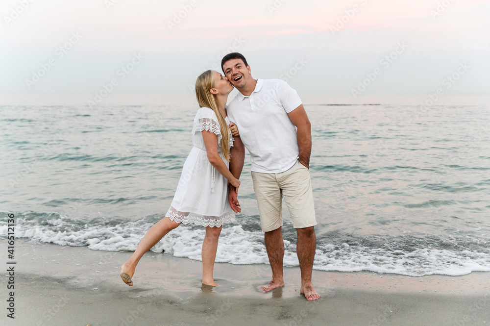 beautiful girl with white hair kisses her husband on the cheek on the beach near the ocean at sunset. romantic walk. vacation of a young family. travel to the sea. beautiful girl with white hair kisses her husband on the cheek on the beach near the ocean at sunset. romantic walk. vacation of a young family. travel to the sea.