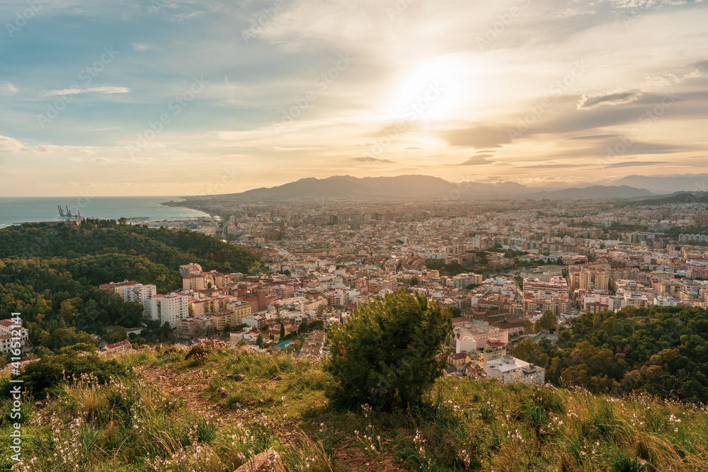 Beautiful panoramic sunset view of the city of Malaga from a viewpoint on a hill with lawn and plants, overlooking the city, the sea, the mountains and the cloudy sky in the background.