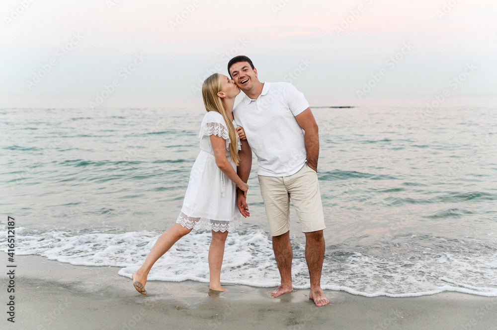 beautiful girl with white hair kisses her husband on the cheek on the beach near the ocean at sunset. romantic walk. vacation of a young family. travel to the sea.