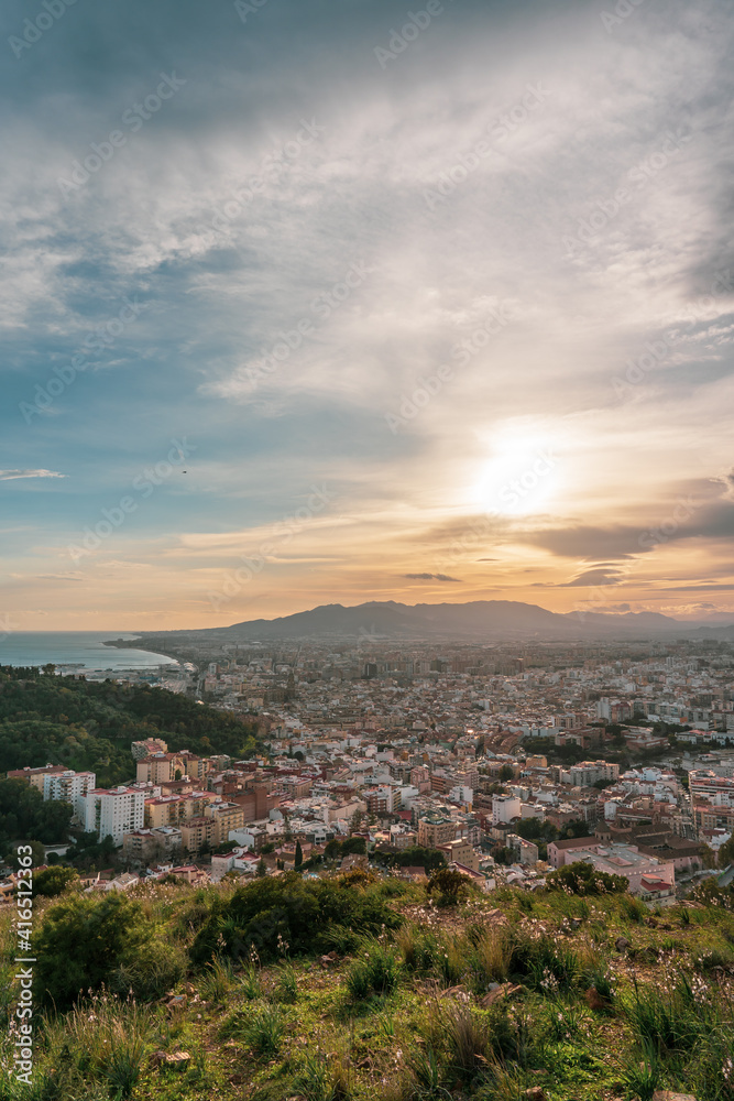 Beautiful sunset views of the city of Malaga from a viewpoint on a hill, overlooking the city, the sea, the mountains and the cloudy sky in the background.