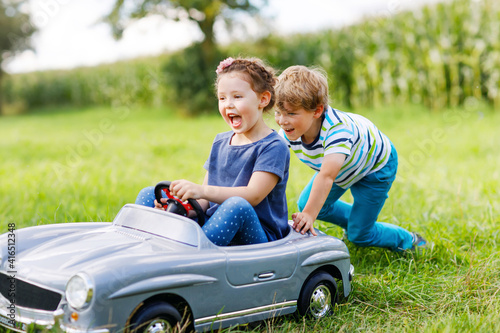 Two happy children playing with big old toy car in summer garden, outdoors. Boy driving car with little girl inside. Laughing and smiling kids. Family, childhood, lifestyle concept © Irina Schmidt