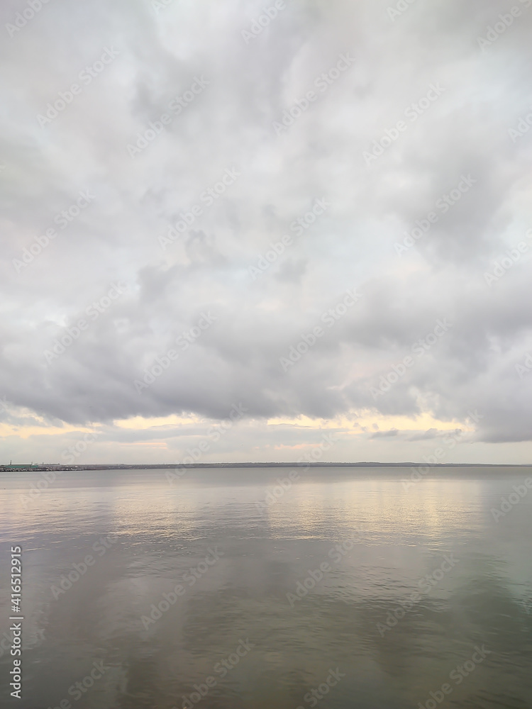 The water surface of the reservoir on a rainy cloudy day. Landscape in dark gray tones. Cumulus clouds hang over the bay. Sunlight and rays barely break through. Vertical photography.