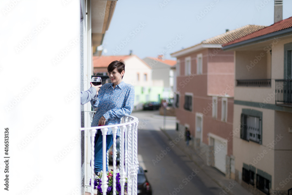 Una mujer joven sujeta una copa de vino tinto en el balcón y brinda con otra persona que saca el brazo desde el interior de la casa