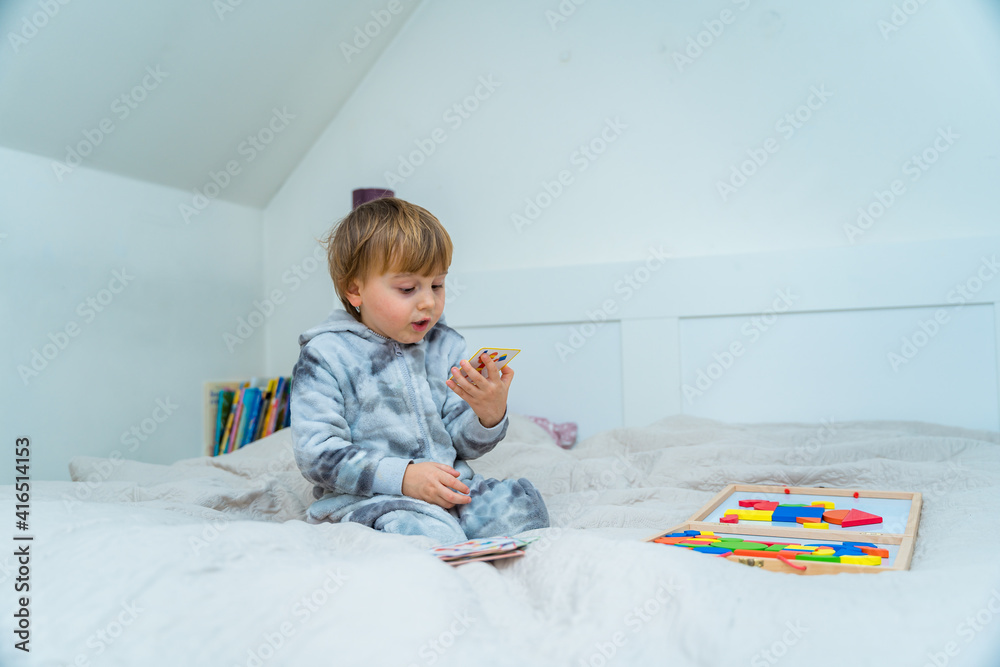 Little toddler boy plays on the bed in a wooden magnetic constructor, preschool child learning at home to make mosaic. Early development. Playing with chikldren during coronavirus outbreak. Little toddler boy plays on the bed in a wooden magnetic constructor, preschool child learning at home to make mosaic. Early development. Playing with chikldren during coronavirus outbreak.
