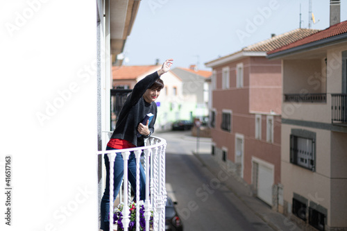 Una mujer joven que sujeta un libro con una mano y saluda con la otra desde el balcón 