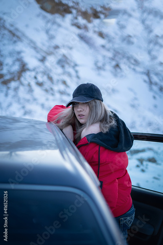 A beautiful girl in a red jacket and black baseball cap with her hair flying in the wind leaned against the car and bared her shoulder sexually. In winter, against the background of snow mountains