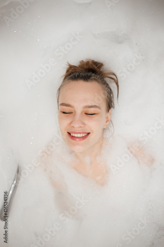 Beautiful portrait of young happy smiling woman takes a bath all in foam. Spa relaxation and self care	
