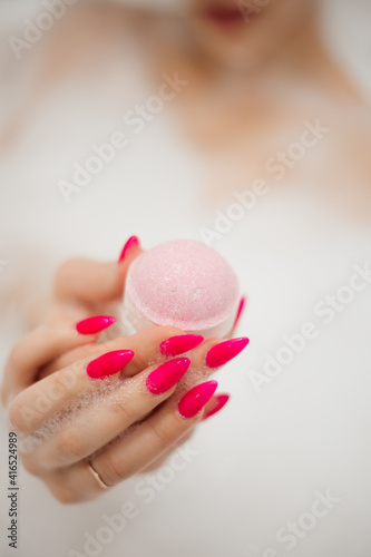 Female hand with a bright pink manicure holds a bath bomb in the foam