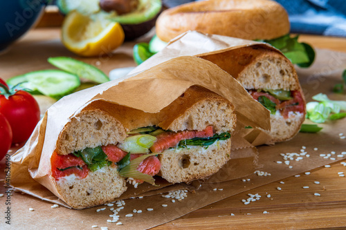 Stuffed toasted bagel sandwich cut into half with smoked salmon, cream cheese, cucumber lettuce. Avocado bagel in the background on a baking sheet. Gluten free healthy breakfast, vibrant fun colors 