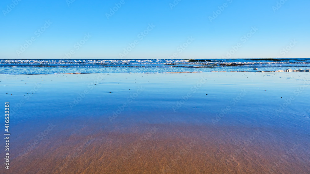 Gentle waves breaking against the shores of a sandy beach with cloudless blue sky, surf, and blue sky reflections in wet sand