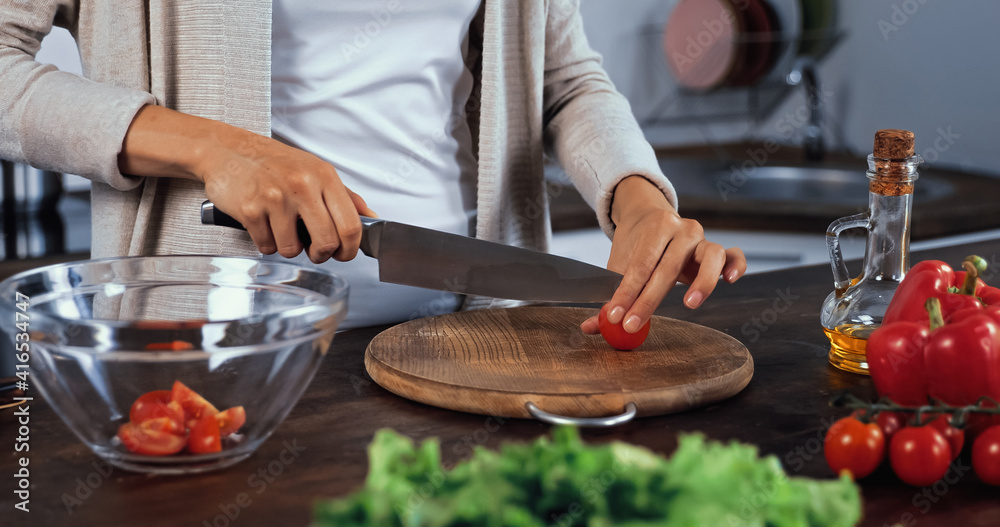 Cropped view of woman cutting cherry tomato near oil and vegetables on blurred foreground Cropped view of woman cutting cherry tomato near oil and vegetables on blurred foreground