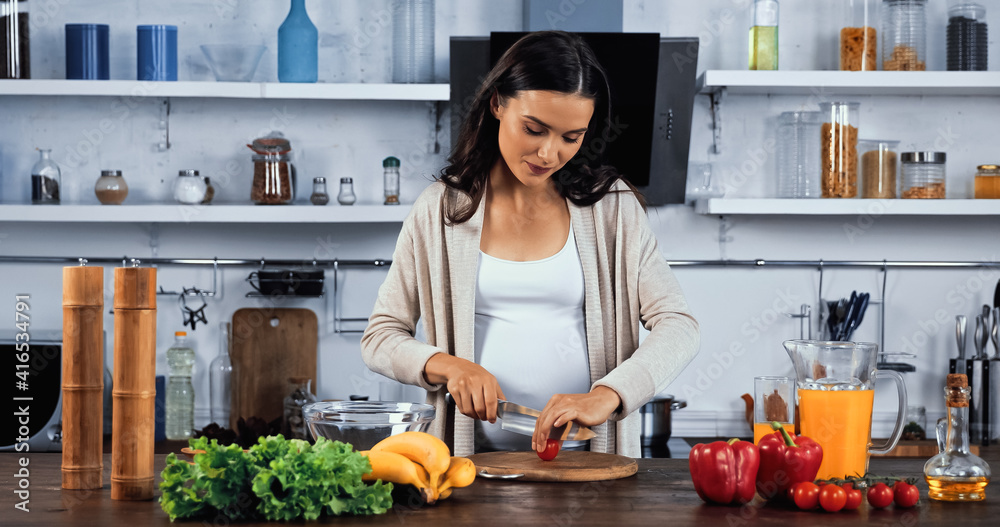 Pregnant woman cutting cherry tomato near fresh ingredients on kitchen table Pregnant woman cutting cherry tomato near fresh ingredients on kitchen table