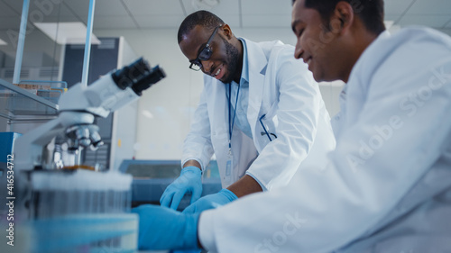 Modern Medical Research Laboratory: Two Smiling Male Scientists Working Together Using Microscope, Analysing Samples, Talking. Advanced Scientific Lab for Medicine, Biotechnology.