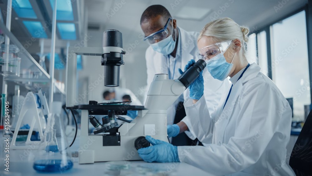 Modern Medical Research Laboratory: Two Scientists Wearing Face Masks Working Together Using Microscope, Analysing Samples, Talking. Advanced Scientific Lab for Medicine, Biotechnology. Modern Medical Research Laboratory: Two Scientists Wearing Face Masks Working Together Using Microscope, Analysing Samples, Talking. Advanced Scientific Lab for Medicine, Biotechnology.