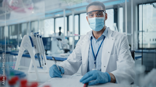 Medical Research Laboratory  Portrait of Male Scientist Wearing Face Mask Looking at Camera  Writing Down Information. Advanced Scientific Lab for Medicine  Biotechnology  Microbiology Development