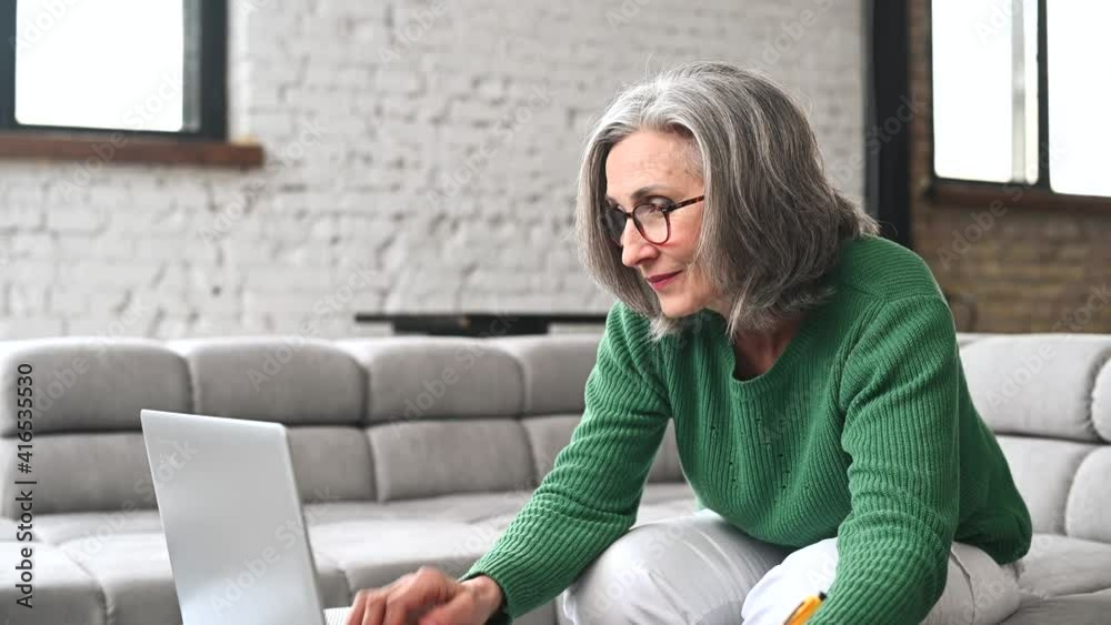 Charming senior online teacher conducts webinar, online classes, mature modern elderly woman sitting with a laptop at home, studying, taking a notes. An elderly modern business lady works remotely