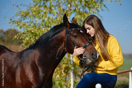 Young female farmer taking care about horse on ranch on summer day, copy space. Girl stroking and hugging brown horse