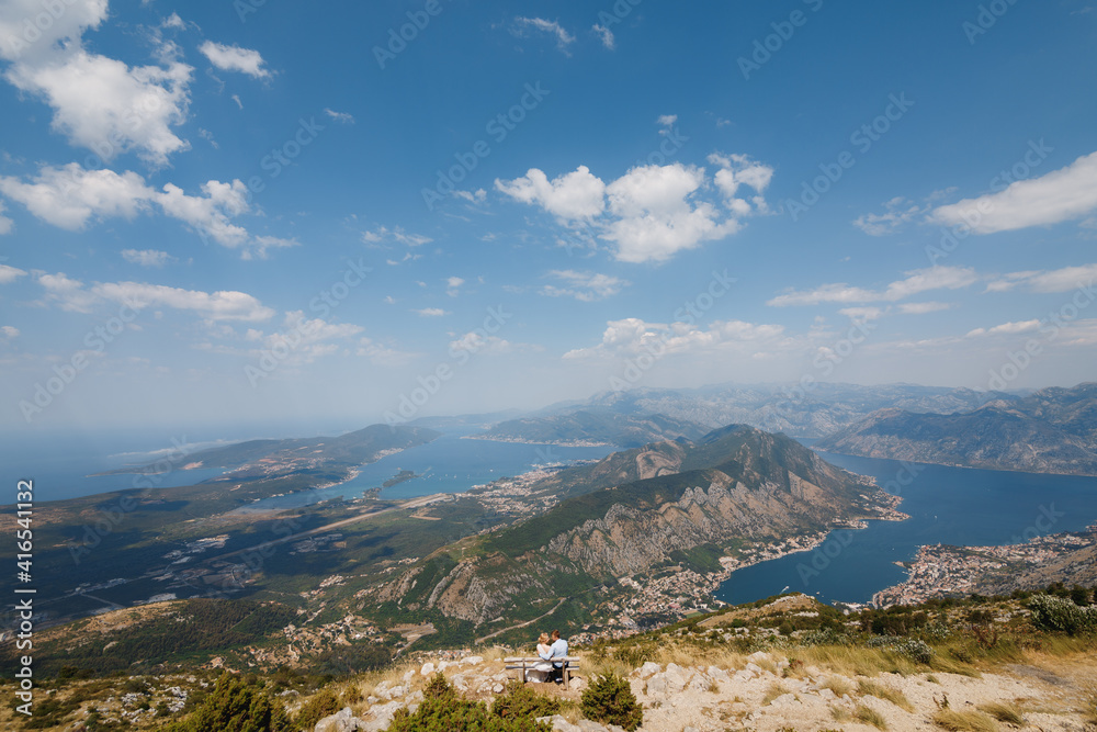 A man and a woman are sitting in an embrace on a bench, a panoramic view of the Bay of Kotor opens in front of them 