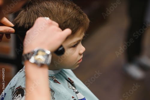 A little cute boy sits in a hairdresser's at the stylist's, a schoolchild is getting hair cut in a beauty salon, a child at a barbershop's, a short men's haircut.