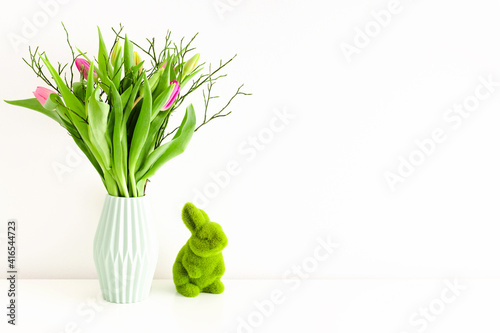 Bouquet of multicolored unopened tulips in light green ceramic vase and Easter green grass bunny on white wall background. Holiday decoration concept. Place for text.