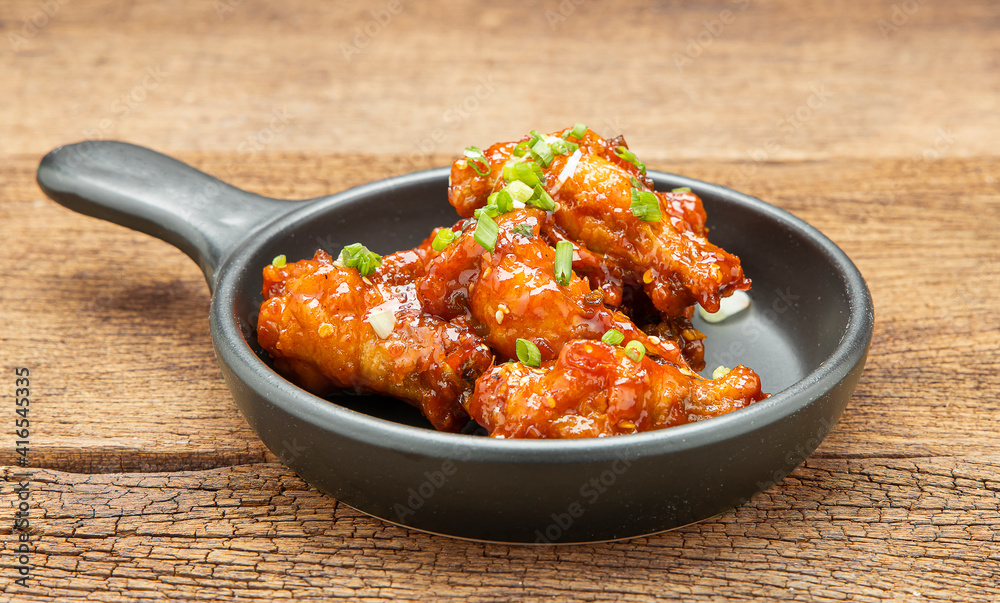Chicken stir-fried with spicy sauce sprinkled with white sesame and chopped scallions is placed in a black ceramic pan on a wooden table. Chicken stir-fried with spicy sauce sprinkled with white sesame and chopped scallions is placed in a black ceramic pan on a wooden table.