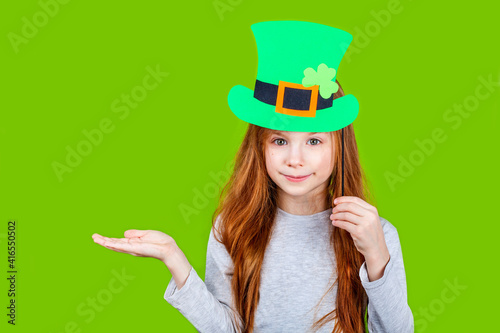 Little smiling caucasian red-haired girl in green hat of leprechaun for irish St. Patrick's Day on green studio background. Copyspace.