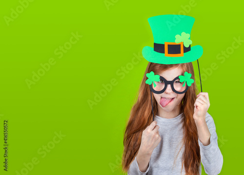 Little smiling caucasian red-haired girl in green hat of leprechaun and shamrock clover glasses sticking out tongue Irish St. Patrick's Day. Copyspace.