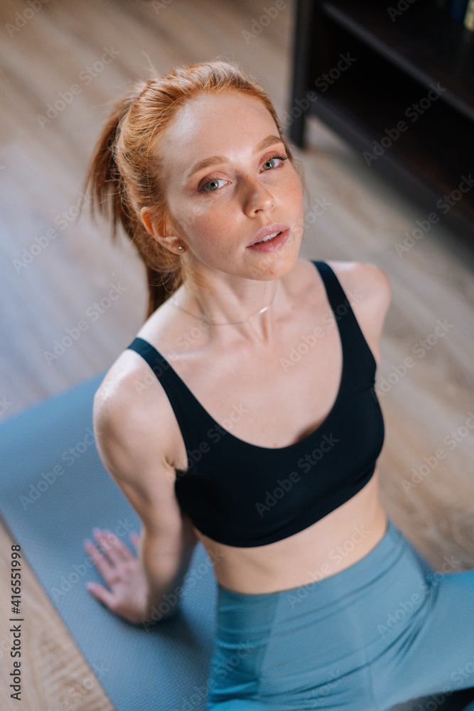 High-angle view of sporty redhead young woman wearing sportswear sitting in floor on yoga mat, looking at camera. Concept of sports training red-haired lady during quarantine.