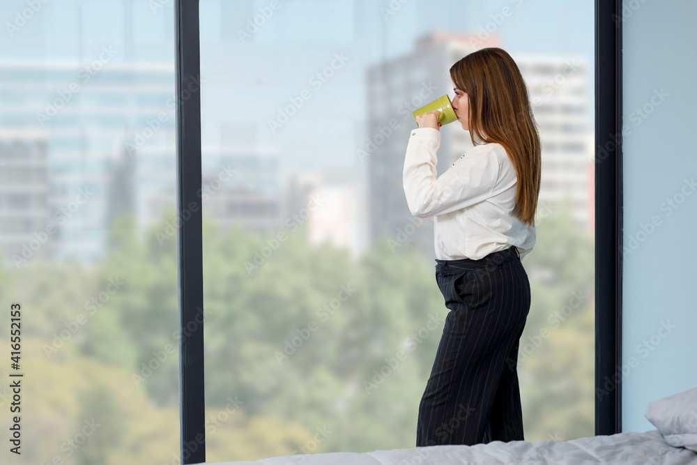 Fototapeta premium Mujer tomando café mientras mira por la ventana de su recamara. Concepto de actividades en casa.