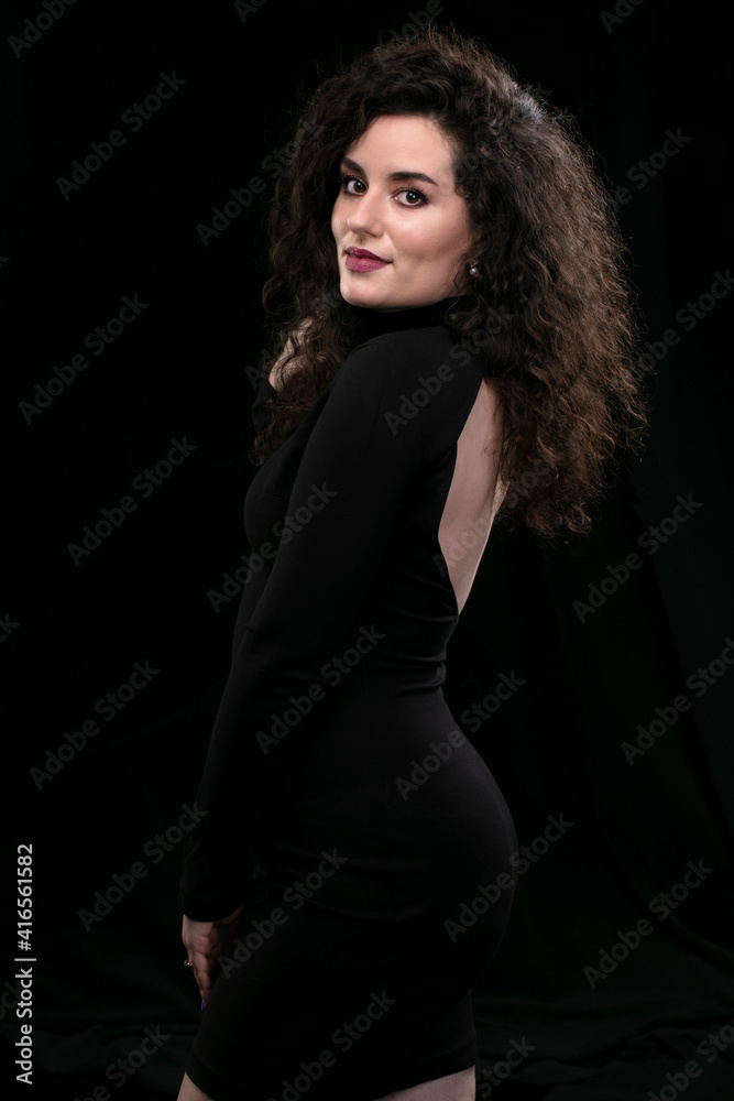 Beautiful brunette girl with curly hair in a tight black dress on a dark background. Beautiful brunette girl with curly hair in a tight black dress on a dark background.