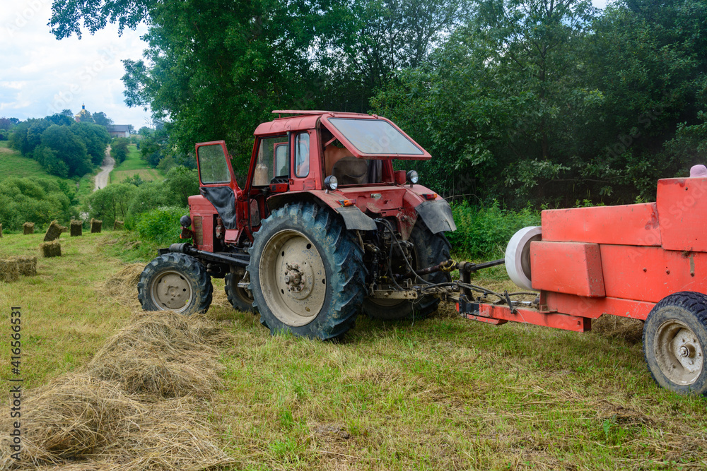 Old red tractor in the field during the haymaking season, pressing hay on bales, forage harvesting. Old red tractor in the field during the haymaking season, pressing hay on bales, forage harvesting.