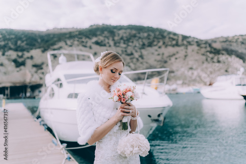 A beautiful bride in a vintage dress with a wedding bouquet and a clutch in her hand poses against the background of yachts at the pier. © manifeesto