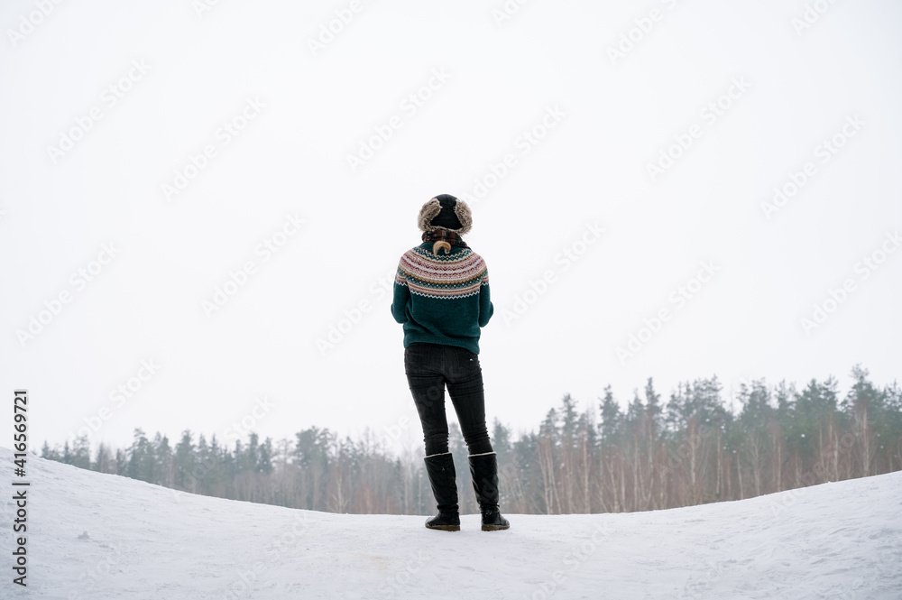 A girl in a knitted sweater and a fur hat stands on snow with her back to the camera. Scenic winter landscape. Beautiful forest.