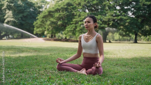 Asian young woman sitting on the grass in the lotus position and raising hands up outside in city park with the big trees background. Rearview of female practicing yoga outdoors on a sunny day.