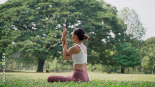 Back view of an unrecognizable slender Asian young woman sitting on the grass in the lotus position and raising hands up outside in city park with the big trees background.