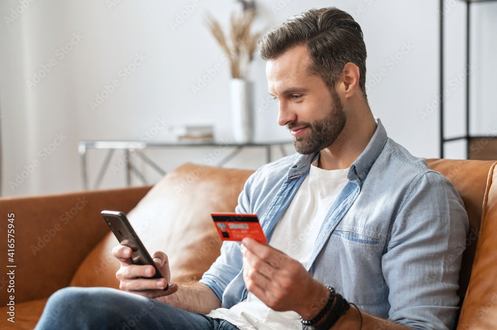 Bearded hipster guy businessman, holding mobile phone and credit card for online shopping and entering number to make payment, paying for utilities, using a virtual cabinet to manage banking account Bearded hipster guy businessman, holding mobile phone and credit card for online shopping and entering number to make payment, paying for utilities, using a virtual cabinet to manage banking account