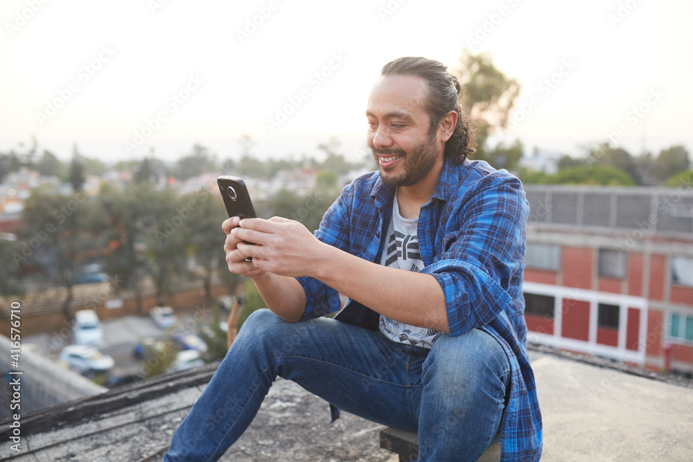 30-year-old man sitting on his back on the rooftop of a building watching the sunset and browsing on his smart cell phone 30-year-old man sitting on his back on the rooftop of a building watching the sunset and browsing on his smart cell phone