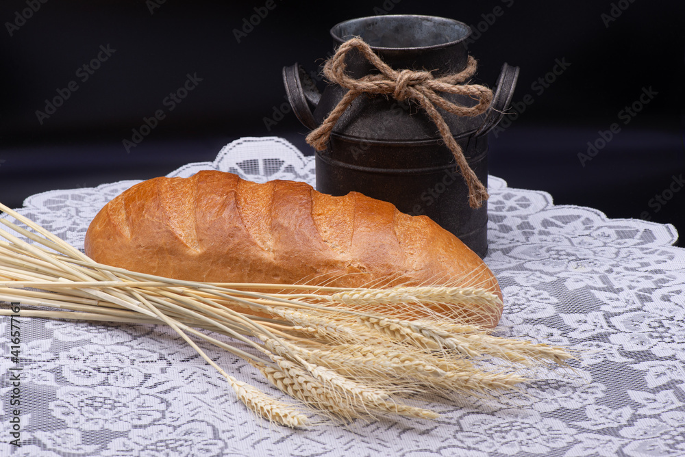 Composition of white bread, wheat, an old jug on white tablecloth on black background