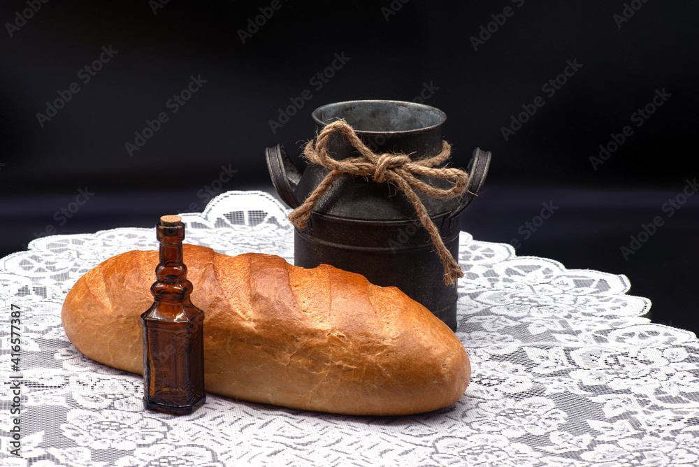 Composition of white bread, small bottle and old jug on white tablecloth on black background
