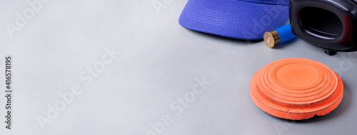 Skeet shooting. Clay target, noise cancelling headphones, bullet shell, and blue cap on gray. Wide banner with copy space