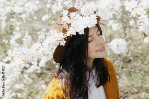 Stylish beautiful woman in hat sensually posing in blooming cherry branches in sunny spring, happy
