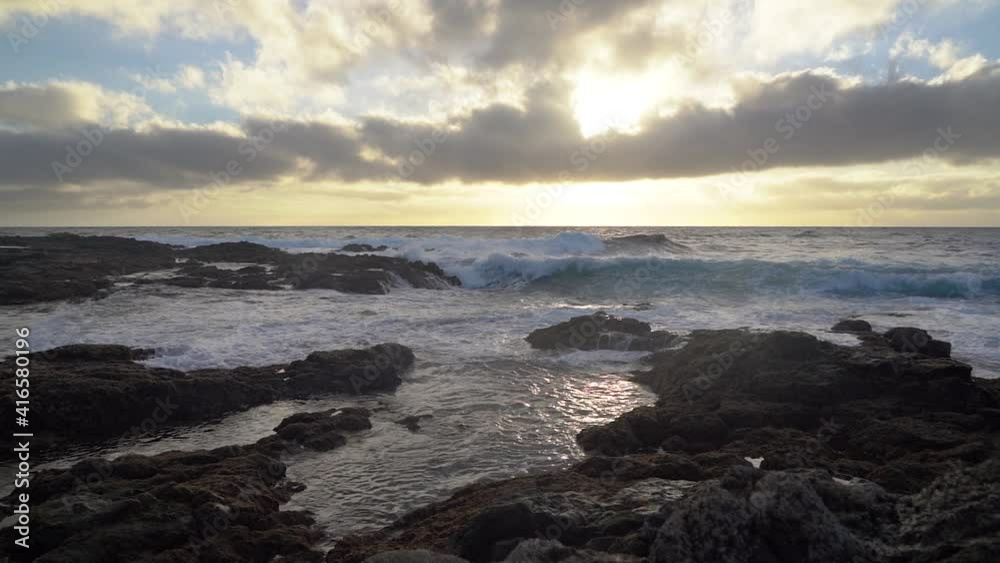 slow motion sunset in seascape with waves crashing on the rocks and sun setting on the horizon.