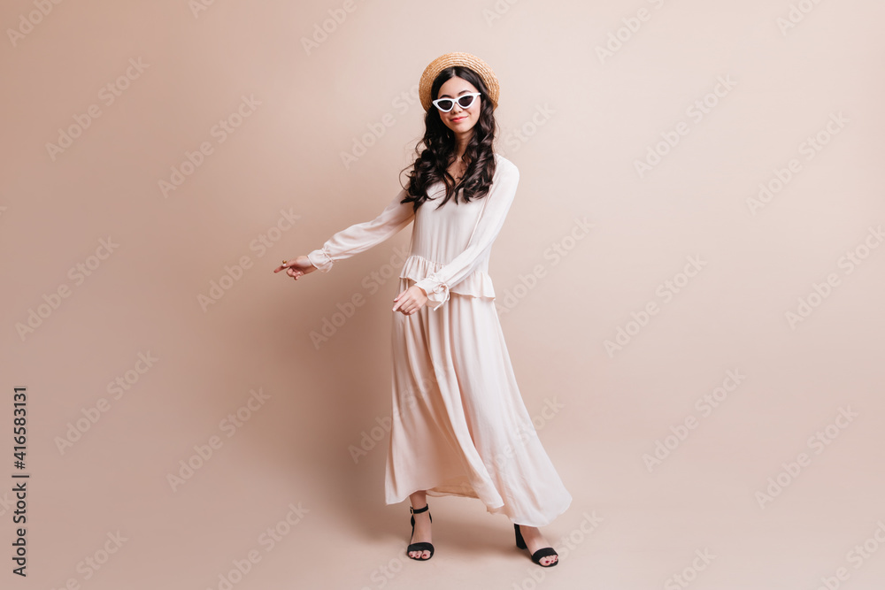 Full length view of elegant asian woman in hat. Studio shot of fashionable japanese girl standing on beige background. Full length view of elegant asian woman in hat. Studio shot of fashionable japanese girl standing on beige background.