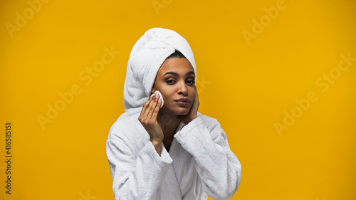 African american woman in towel and bathrobe cleaning face with cotton pad isolated on yellow © LIGHTFIELD STUDIOS