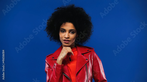 Curly african american woman in red jacket looking at camera isolated on blue