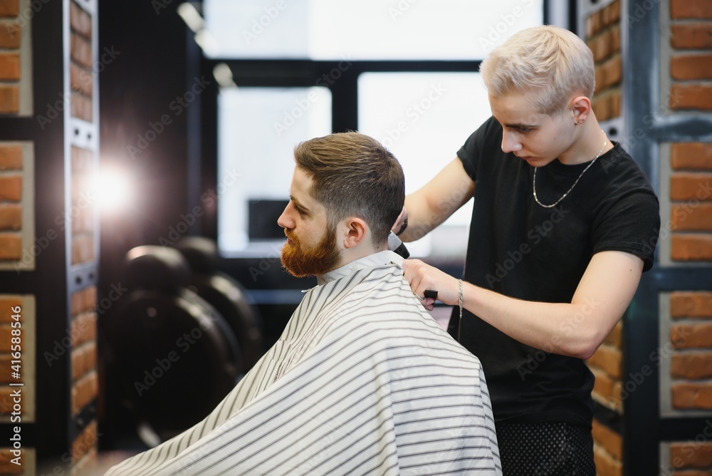 Obraz premium Making haircut look perfect. Young bearded man getting haircut by hairdresser while sitting in chair at barbershop