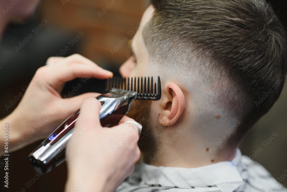 Making haircut look perfect. Young bearded man getting haircut by hairdresser while sitting in chair at barbershop